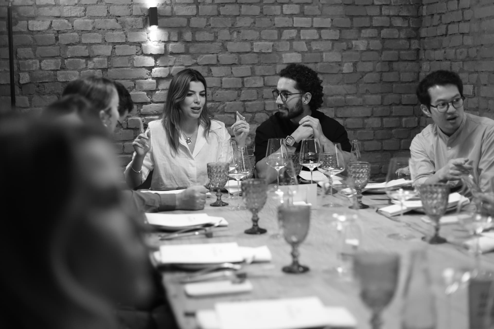 Convidados em conversa ao redor de uma mesa longa em sala de tijolos